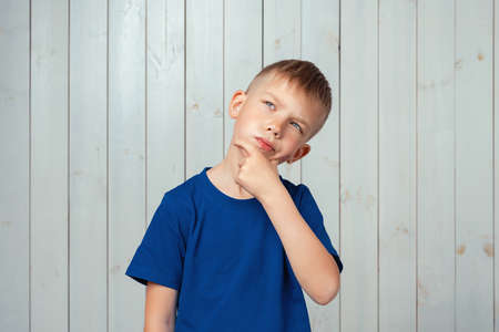 Portrait of dreamy preteen boy in blue t shirt daydreaming about having something, touching chin and pondering. Studio shot, light wooden backgroundの写真素材