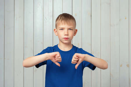 Serious preteen boy in blue t shirt showing thumb down and expressing dislike. Studio shot, light wooden backgroundの写真素材