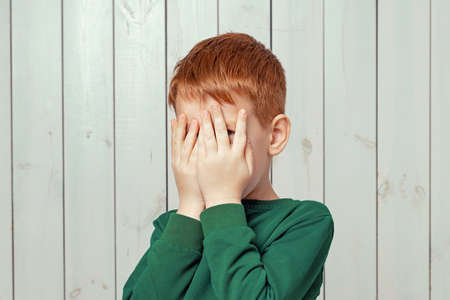 Close up scared ginger little boy 7-9 years hiding face in palms hands and peering out with one eye...