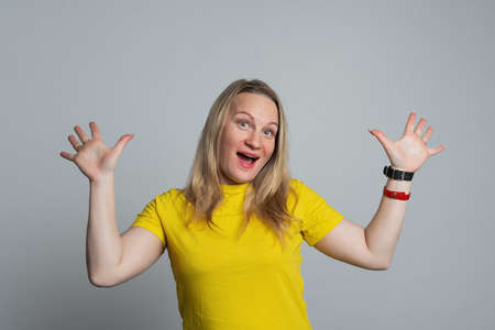 Surprised happy blonde woman wearing in casual yellow t shirt rejoices and spreads her hands to the...