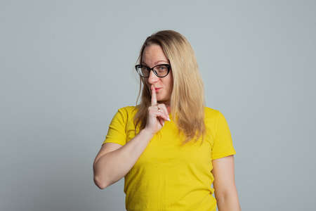 Portrait of mature woman in glasses keeping finger at her mouth, saying Shh, Hush, Tsss, wearing...