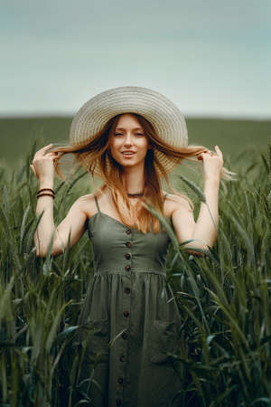 Young woman in a hat walking in a wheat field, enjoys life and summer. Wheat field. Beautiful Nature Landscape under Sunlight. Healthy lifestyle Concept. Image with noise, film grainの写真素材