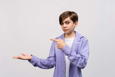 Handsome boy 12-14 years old looking at the camera and showing aside on copyspace. Pay attention to important information. Studio shot on light gray backgroundの写真素材