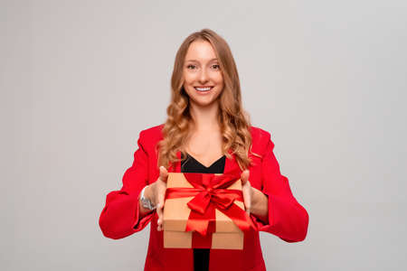 Smiling young blonde woman holding gift box decorated with red bow. Attractive girl in stylish red jacket posing isolated on gray background. New Year Women's Day birthday holiday conceptの写真素材