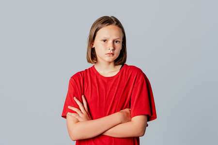 Serious-looking offended teen girl in casual red t shirt, sulking frowning upset, cross arms chest block unsatisfied pose, boy insulted her waiting apology complaining displeased, stand gray backgroundの写真素材