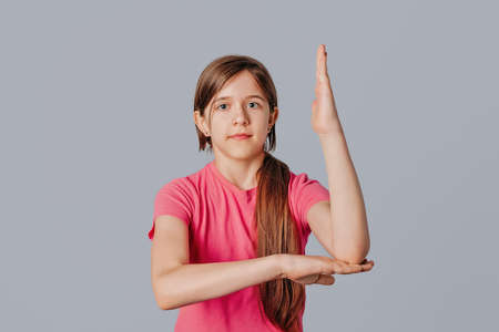 School girl in casual pink t shirt raising hand up, standing over gray background. Teen girl ready to ask teachers question. Can be used for topics like education, studying, schoolの写真素材