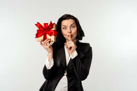 Happy businesswoman in black formal suit jacket posing on white background with gift box decorated with red ribbon. Holidays, celebration and lifestyle conceptの写真素材