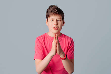 Teenager boy in casual pink t shirt hold hands in prayer hope for luck, isolated over gray color backgroundの写真素材
