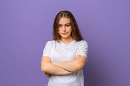 Portrait of upset brunette woman in white casual t shirt standing with arms folded over purple...