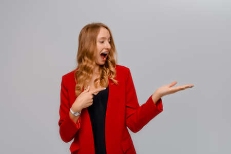 Portrait of smiling excited blonde young woman pointing at something interesting, looking happy at product, standing in red blazer against gray backgroundの写真素材