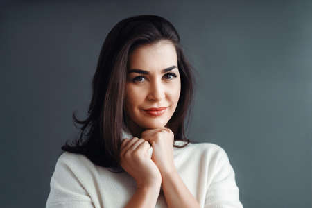 A beautiful brunette woman in a white sweater poses on a gray background in the studio. Beauty and elegance.の写真素材