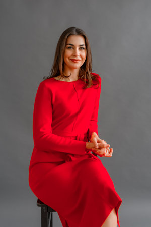 Carefree young woman in red dress sitting on a chair, posing in studio over gray background with a happy smile on face. Relaxed and beautiful young womanの写真素材