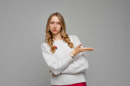 Disappointed blonde young woman pointing hand right, looking upset and frowning skeptical, showing at promo offer, stands in white sweater against gray backgroundの写真素材