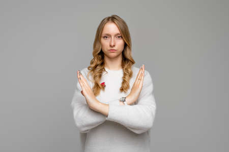 Serious blonde young woman wears white sweater showing stop gesture with crossed hands, trying to defend herself as if saying: Stay away from me. stay home. social distancing. New normal behaviorの写真素材