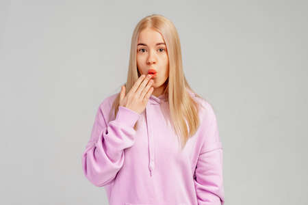 Portrait of blonde girl with guilty big eyes looks confused, cover mouth lips hand on mouth, wears violet hoody, stands against gray background. Oops, oh, ouch gestureの写真素材