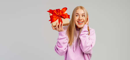 Portrait of Happy young blonde woman in trendy hoodie holding gift box with red ribbon and looking...