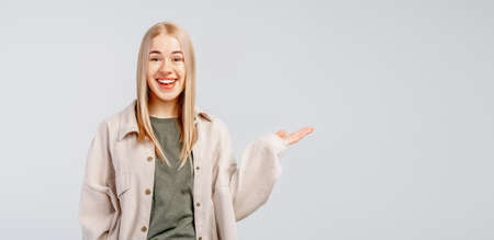 Happy smiling stylish blond girl in trendy shirt show something in hand, standing over gray...