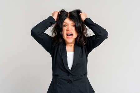 Portrait of young dark-haired girl in panic shouting and grabbing her hair in fear or frustration, standing in black blazer over gray background. stress and mental healthの写真素材