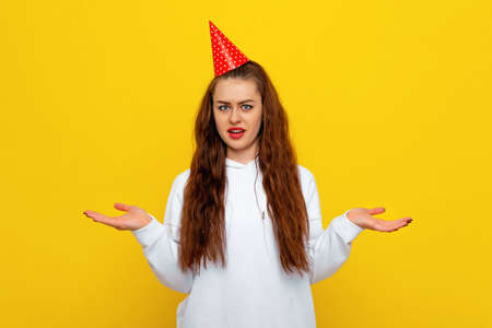 Shocked perplexed confused young woman in white casual style sweatshirt and birthday hat shrugs shoulders with hesitation, faces doubtful situation, spreads hands, hard decisionの写真素材