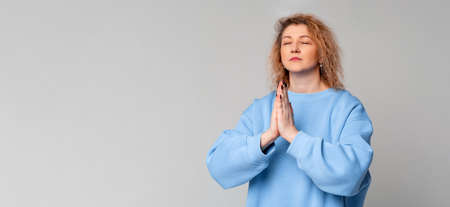 Grateful hopeful middle age woman stands in meditative pose, enjoys peaceful atmosphere, holds hands in praying gesture, has sense of inner peace, standing over gray background. meditation conceptの写真素材