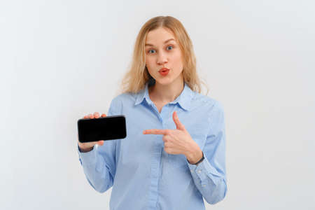 Amazed blond girl looking at camera and pointing finger at mobile screen, recommend online shopping store, horizontal smartphone, application interface. Indoor studio shot on white backgroundの写真素材