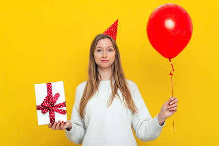Pretty young woman in a birthday cap and white casual style sweater holds in hands box with gift and red inflated helium balloon, celebrates birthday or special occasion. holiday and party conceptの写真素材