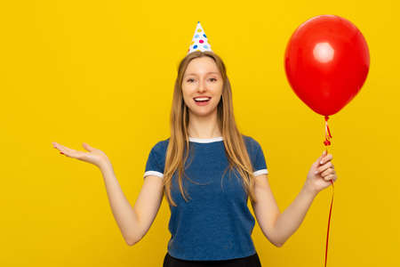 Pretty smiling young woman in a birthday cap gestures with hand, holds red inflated helium balloon, celebrates birthday or special occasion. holiday and party conceptの写真素材