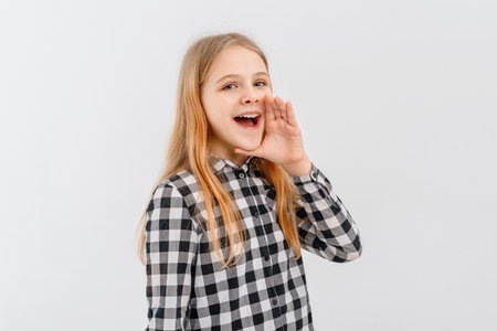 Attractive teen girl holding palm near wide open mouth is announcing, telling a secret, shouting or yelling, standing in casual plaid shirt over white background. Communication and promo conceptの写真素材