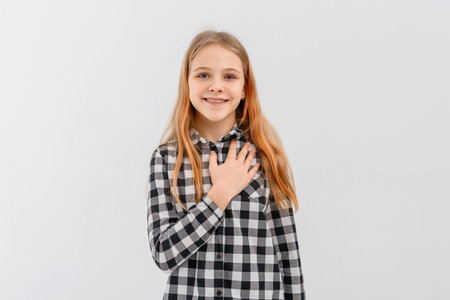 Smiling teen girl looks flattered, holds hand on chest, feels appreciation, stands in casual plaid shirt over white studio backgroundの写真素材