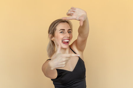 Young lovely woman wearing casual black top, stand over beige studio background, smiling making frame with hands and fingers with happy faceの写真素材