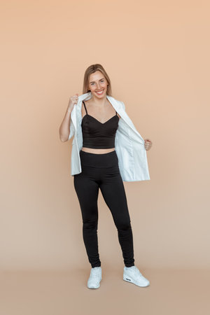 Full length of smiling young woman with freckles posing in white blazer and black clothes with carefree upbeat face expression. Indoor studio shot on beige backgroundの写真素材