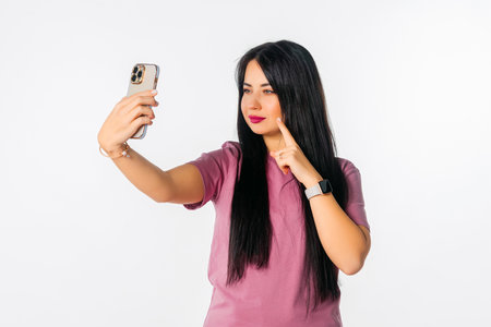 Serious young woman holding smartphone using mobile phone app for video call, standing against white studio backgroundの写真素材