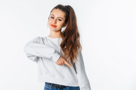 Happy young woman in white sweatshirt, pointing finger down, showing amazing news, promo banner, offer. Smiling brunette female stands against white studio backgroundの写真素材