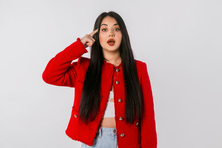 Young woman having a great idea. Portrait of young girl with long hair, pointing finger at the temple, standing in red blazer against white studio backgroundの写真素材