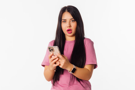Amazed girl using mobile phone, holding telephone, looks impressed, stands over white background. Pretty brunette female holding cell phone and reading funny message she received from boyfriendの写真素材