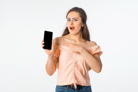 Surprised young woman in stylish outfit, pointing at mobile phone screen, showing smartphone display and looks amazed and impressed by smth, white backgroundの写真素材