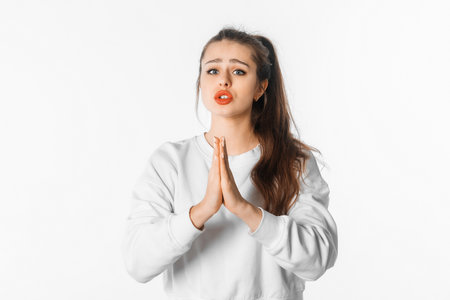 Beautiful young woman in casual clothes holding hands together and praying, posing against white studio backgroundの写真素材