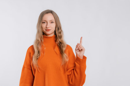Cheerful attractive young woman points finger up, shows way to banner, demonstrates logo or advertisement on top, stands in an soft orange sweater over neutral backgroundの写真素材
