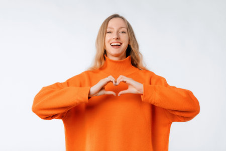 Lovely young woman shows heart sign, i love you gesture express her care and sympathy, stands in soft orange sweater over neutral studio backgroundの写真素材