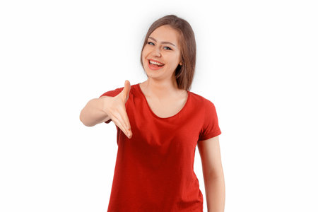 Young friendly woman smiling cheerful, offering a handshake as greeting and welcoming. Winsome female model in red t shirt posing over white studio backgroundの写真素材