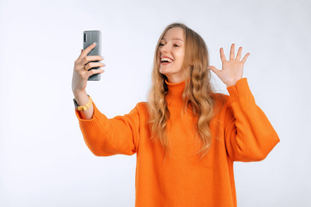 Joyful young woman wearing soft orange sweater doing selfie shot on mobile phone waving greeting with hand standing over neutral background, studio portraitの写真素材