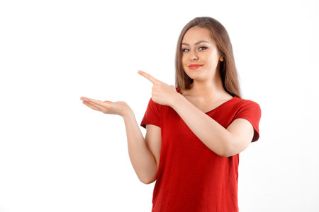 Surprised lovely girl points left at promo banner, shows advertisement on blank white space. Young brunette female looking amazed by promotion offer, stands over white backgroundの写真素材