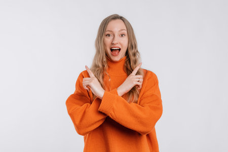 Positive friendly young woman with a happy smile on face pointing fingers sideways, showing left and right promo, stands in an soft orange sweater over neutral background. Great idea cool offerの写真素材