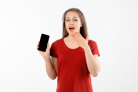 Amazed girl show smartphone, looks impressed, stands over white background. Pretty brunette female holding cell phone with blank screenの写真素材