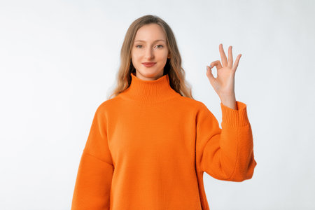 Portrait of beautiful happy woman showing okay sign, stands in soft orange sweater over neutral backgroundの写真素材