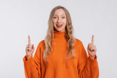 Positive young woman points fingers up, smiles pleasantly, stands in an soft orange sweater over neutral background. Great idea cool offerの写真素材