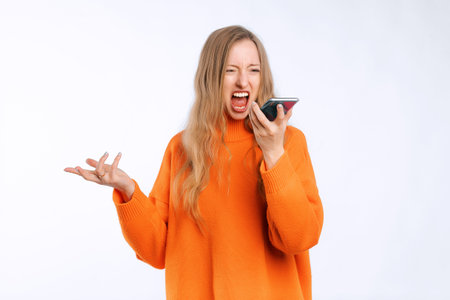 Angry woman arguing on call, screaming at mobile phone speakerphone, recording voice message with furious face, standing in an soft orange sweater over white backgroundの写真素材