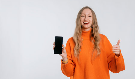 Smiling lovely woman shows a mobile phone with a blank screen, stands in an soft orange sweater over neutral backgroundの写真素材