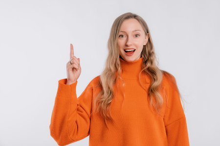 Smiling attractive young woman points finger up, shows way to banner, demonstrates logo or advertisement on top, stands in an soft orange sweater over white backgroundの写真素材
