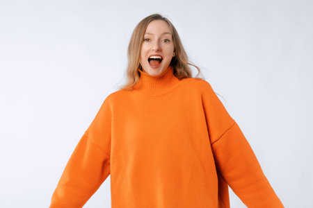 Portrait of smiling cheerful girl, looks happy and upbeat at camera, stands in trendy knitwear orange sweater against neutral studio backgroundの写真素材